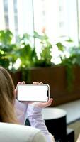 sostener un de cerca teléfono con un blanco pantalla espacio para texto un niña se sienta cerca el ventana con plantas en el antecedentes con su espalda a el pantalla en su manos sostener un artilugio video