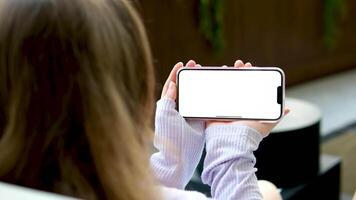 houden een detailopname telefoon met een wit scherm ruimte voor tekst een meisje zit in de buurt de venster met planten in de achtergrond met haar terug naar de scherm in haar handen houden een apparaatje video