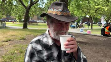 hasting street vancouver elderly man with gray beard drinks tea on the street with a plastic cup paper cup cowboy hat plaid shirt homeless people tea party hot drinks help drug legalization video