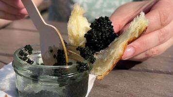 with a glass jar a wooden spoon scoop black caviar on a bun on a croissant tasting tasting delicious food caviar fish lumpfish with a wooden table close-up hands on the street picnic rich life video