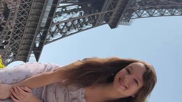 joven hermosa niña en un azul vestir se sienta cerca el eiffel torre cerca un verde árbol ella endereza su trenzas mira alrededor ella es contento y muy contento video