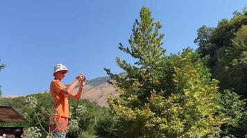 une Jeune gars photographe pousse une prend photographies dans un albanais point de repère bleu œil adolescent contre le Contexte de le ciel et montagnes dans une Panama casquette et un Orange veste video