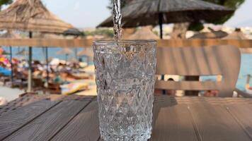 gegen Hintergrund von Strand Regenschirme und Meer, Wasser ist gegossen in Glas schleppend Bewegung hölzern Tabelle im Entfernung das Hintergrund von das Strand ist verschwommen Kühle Hitze Verlangen zu trinken Wasser video