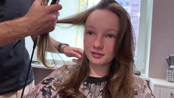 professionnel brunette styliste fait du moderne coiffure Faire permanente dans beauté salon. coiffeur les usages curling le fer sur cheveux de blond client séance près miroir video