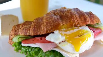 delicioso apetitoso croissant com ovo fluindo gema fluindo Fora do a sanduíche carne tomate salada e brie queijo suco em uma branco prato café da manhã almoço delicioso molho e pepino França Europa video