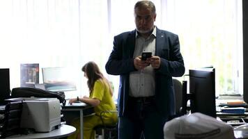 Boss boss in office walks with phone in center behind him, woman girl works computers, fax phone, looking for messages, overweight man in suit in blue jacket against background of the window, busy video