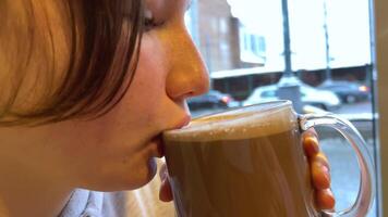 girl face close-up freckles drink hot chocolate cocoa with milk against backdrop of city cars driving winter autumn cold warm up delicious natural product aromatic teenager young real person in cafe video