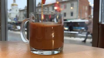 en un café por el ventana remover caliente chocolate. ciudad personas caminando carros conducción Iglesia en el antecedentes allí es nieve frío calentar arriba comodidad sabroso tratar bebida para el nuevo año Navidad vaso vaso video