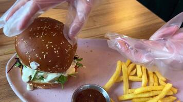 Female hands in gloves form cheeseburger with Brie cheese on a plate with french fries and sauce preparing food serving eating using cellophane plastic transparent gloves convenient in a restaurant. video