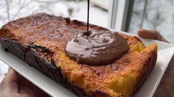 gießen Schokolade Über Kuchen gegen Hintergrund von Fenster cool köstlich Delikatesse Winter, Schnee heiß Käsekuchen Quark Reis Kasserolle Pasta Hütte Käse im öffnen Fenster kalt halt Teller mit Dessert im Hände video