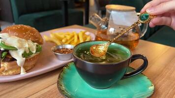 decorative tableware golden tea spoon with emerald stir tea on background burger, cheeseburger, french fries Green cup and saucer lunch, lunch dinner aristocratic food fast food video