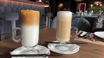 Latte two types cold and hot ice latte and classic on wooden table, milk and coffee serving comparison choice Summer and winter preference close-up two glasses with black straws in cafe. drink layers video