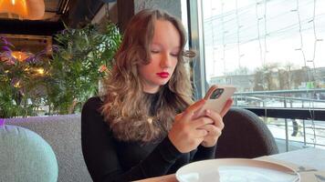 young girl with phone in restaurant bright red lipstick black evening dress write SMS search for wi-fi use Internet play games write on phone notes text sit at table empty plate by window. Christmas video