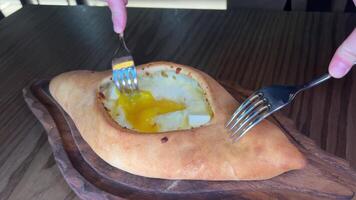 mão mistura ingredientes do adjariano khachapuri com garfo dentro restaurante. aberto pão torta com queijo e ovo gema. gostoso georgiano cozinha. lento movimento. video