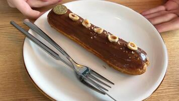 exquisite eclair on white plate with pieces of hazelnut and golden color around knife and fork scroll with hands, looking at delicious dessert, drizzled with milk chocolate, restaurant serving yami video