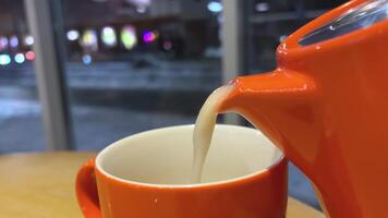 Gießen Tee mit Milch gegen das Hintergrund von ein Nacht Winter Stadt orange, Teekanne Tasse in der Nähe von Fenster Fußgänger London schwarz Tee mit Milch und Zucker Süße zum das Abend köstlich zum ein gut schlafen. video