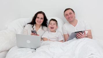 family looks into the frame laughing kid opened his mouth having fun beautiful people with gadgets in their hands in a white bed space for advertising text health treatment joy food medicine video