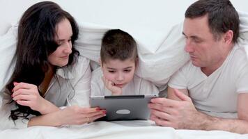 people, family and technology concept mother, father and little boy with tablet pc computer in bed at home or hotel room. Happy family spend time together lying under white blanket on bed at bedroom video