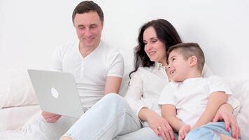 family in bed playing on tablet sincerely laughing having fun spending time together mom father and son dark hair white big duvet family is watching movies with the son on laptop video