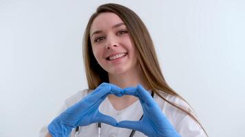 sorridente giovane donna medico cardiologo indossare bianca medico cappotto e stetoscopio mostrando mani cuore forma guardare a telecamera. cardiologia assistenza sanitaria, amore e medicina beneficenza concetto, ritratto. video