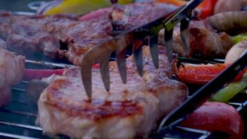 jung kaukasisch Mann flippt heiß saftig Pfeffer mit Zange mit Braten Steak auf das Grill während Stehen im das Hinterhof von seine Haus, Seite Aussicht Nahansicht im schleppend Bewegung mit Zoomen aus. video