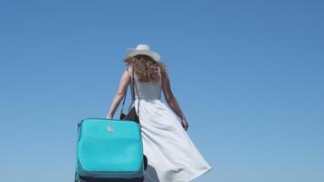 woman girl in white dress tourist with large blue suitcase goes into distance adjusting hat against sky Travel runway place for travel agency advertising text unrecognizable people developing clothes video