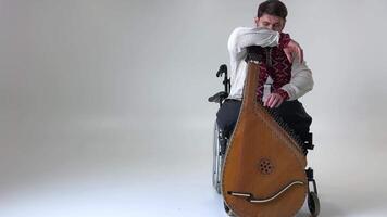 an adult man of 30-45 years old is sitting on a wheelchair holding a musical instrument kobza hand while adjusting it with the other he is wearing Ukrainian vyshyvanka a symbol of Ukraine life music video