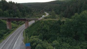 An abandoned rusty railroad bridge above the curving road with a driving truck. Scene. Green summer forest and asphalt bended road under the bridge with supporting columns. video