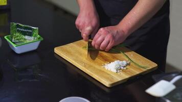 el cocinar es corte verde cebollas. Arte. masculino manos en el marco son corte verde cebollas con cuchillo en el tablero. en el mesa son junta, cebolla, Romero y espátula video