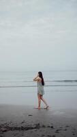 un niña con largo pelo en el Oceano camina en negro arena, vertical video