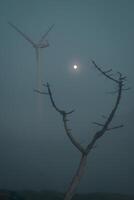 The trunk of a dead pine embraces the full moon with a wind turbine in the background photo