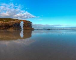 The stone arch of As Catedrais beach is reflected in the waters of low tide photo