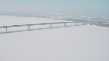 antenne visie van een lang brug bovenstaand sneeuw en ijs gedekt rivier- tussen twee onderdelen van een stad. klem. winter landschap met een breed wit rivier- en een brug met het rijden auto's. video