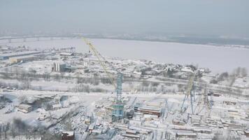 Antenne Aussicht von ein industriell Stadt Zone auf ein Winter Tag und ein breit Eis bedeckt Fluss auf das Hintergrund. Clip. Konstruktion Seite? ˅ mit Kräne und schneebedeckt Stadt Straßen. video