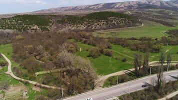 Aerial view of a wide road bending through the green fields covered by trees. Shot. Beautiful valley surrounded by hills and mountains. video