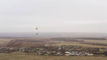 Man with a cloak flying on a paraglider like a Superman over a yellow autumn field. Action. Flying parachutist near the village on a foggy day. video