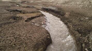 estrecho río fluido y divisor seco suelo. disparo. otoño paisaje con el campo con grietas y frío rápido fluido arroyo. video