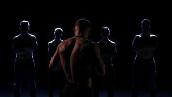 Rear view of a muscular male fighter standing in a dark room against for man silhouettes, unequal confrontation. Stock footage. Four enemies hidden in the shadow, concept of fight with yourself. video