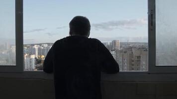 indietro Visualizza di irriconoscibile uomo in piedi su il balcone nel davanti di ha aperto finestra. azione filmato. giovane maschio adolescente guardare a il estate città, vivente su un' alto pavimento. video
