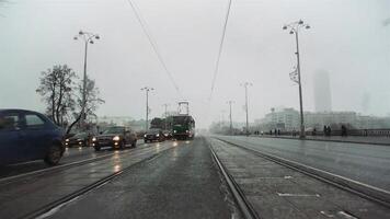 City street with driving cars and trams in late autumn season with heavy fog and falling snow. . Concept of traffic and big city life. video