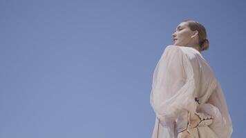 Beautiful woman in white dress on background of sky. Action. Model in stylish dress poses on background of blue sky. Elegant woman in dress on clear day with blue sky video