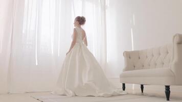 Back of bride in white elegance wedding dress with loop. . Full length. Wedding. Beautiful bride indoors with bouquet of flowers against big window in full lenght back side view video
