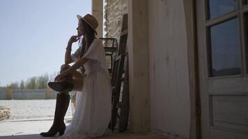 Profile of beautiful young woman in dress. Action. Side view of attractive woman in dress sitting in beautiful pose on chair. Young woman in cowboy style poses sitting on veranda video