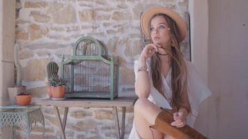 Beautiful young female model in western style sitting in front of brick wall, cactus, and an empty birdcage. Action. A woman with many braids wearing cowboy boots, a hat, and a white dress. video
