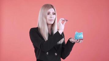 joven hembra poniendo un moneda en cerdito banco, ahorro dinero concepto. acortar. negocio rubio mujer posando a el foto estudio con un azul cerámico cerdo banco y un moneda. video