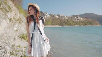 joven hembra adolescente modelo posando por el mar y montaña y disfrutando verano. acción. mujer con Rizado pelo vistiendo blanco vestir y un sombrero en pie por el mar costa. video