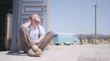 un hombre modelo relajante y sentado en un polvoriento la carretera en un caliente verano día. acción. hermoso hombre en frente de azul puerta, cielo, y pequeño autobús para de viaje. video