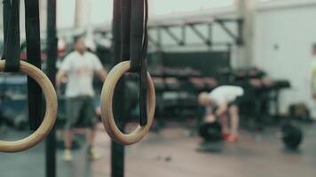 Professional sports equipment and the interior of a gym. . Close up of gymnastic rings with a sports hall with two men lifting heavy weights on the background. video