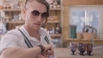 Portrait of a young male model sitting inside cafe in sunglasses at the wooden bar counter. Action. Young man taking out an earphone and looking tired. video