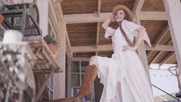 país niña en el espalda porche, blanco vestir y vaquero sombrero. acción. fondo ver de un posando hembra modelo con su pierna en marrón vaquero bota en un de madera mesa. video